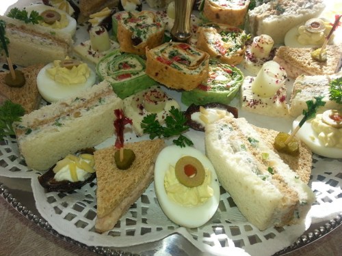 A platter of assorted finger sandwiches, rolled wraps, deviled eggs, and garnishes, elegantly arranged for a gathering.