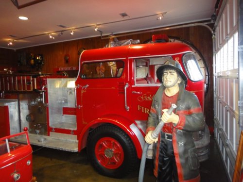 A vintage red fire truck parked in a garage next to a life-sized firefighter statue holding a hose.