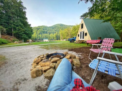 A cozy campsite with a fire pit, chairs, and a cabin surrounded by green hills and trees.
