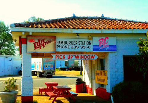 A small hamburger stand with a pick-up window, featuring signs for Dr. Pepper and RC Cola, and red picnic tables outside.