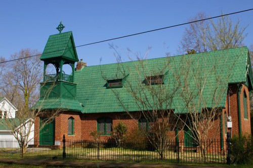 A brick building with a green roof and bell tower, surrounded by trees and a fence, under a clear blue sky.