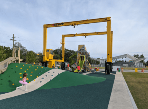 A colorful playground featuring climbing structures and large yellow cranes in the background under a cloudy sky.