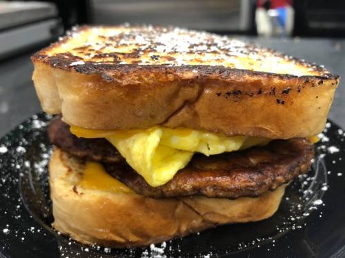 A close-up of a sandwich with toasted bread, cheese, egg, and sausage, garnished with powdered sugar.