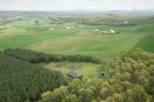 Aerial view of lush green fields, scattered farms, and a forested area under a cloudy sky.