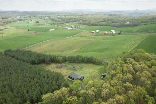 Aerial view of lush green fields, scattered farms, and a forested area under a cloudy sky.