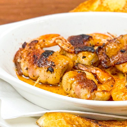 A dish of grilled shrimp and bacon, served in a white bowl with a golden-brown toast on the side.
