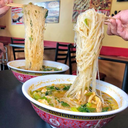 Two bowls of noodle soup with long, thin noodles and vegetables, being lifted with chopsticks.
