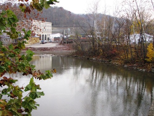 A calm river reflects trees and a building, surrounded by autumn foliage and distant hills.