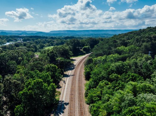 Aerial view of a winding railway track surrounded by lush green trees and distant mountains under a blue sky.