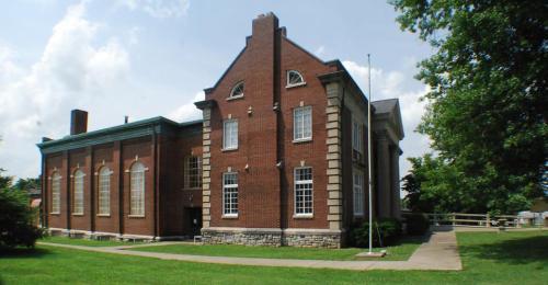 Historic brick building with large windows, set in a green landscape under a partly cloudy sky.