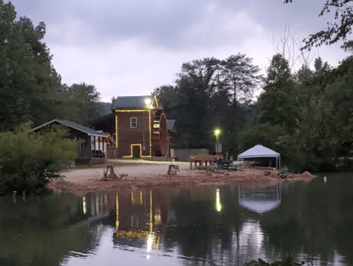 A rustic building with a waterwheel beside a calm lake, surrounded by trees under a cloudy sky. Soft lights illuminate the scene.