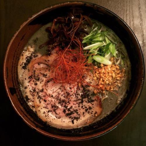 A bowl of ramen with dark broth, sliced pork, green onions, seaweed, and red chili threads on top.