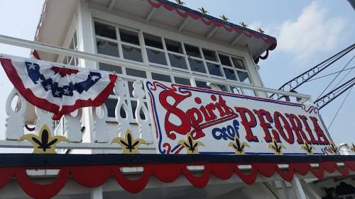 Sign on a boat named "Spirit of Peoria," decorated with red, white, and blue bunting against a clear sky.
