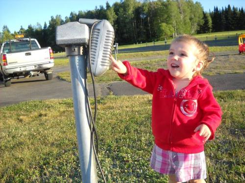 A young girl in a red jacket smiles while reaching for a vintage drive-in speaker in a grassy area.