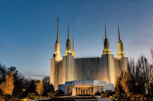 A grand building with tall spires illuminated at dusk, surrounded by trees and decorative lights.