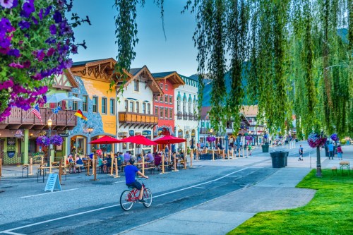 A charming street lined with colorful buildings, outdoor dining, and hanging flower baskets, with a cyclist passing by.