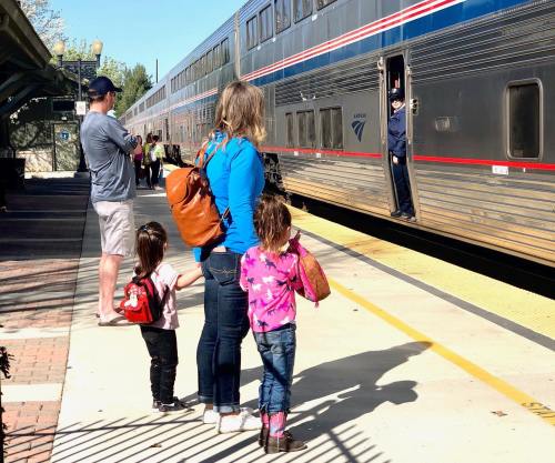 A family waits at a train station as a conductor stands by an Amtrak train. Two children watch eagerly.