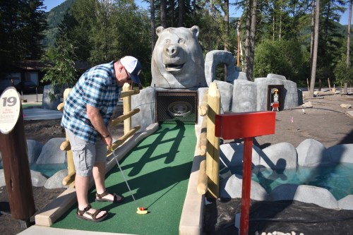 A man plays mini-golf on a green course with a large bear sculpture in the background, surrounded by trees.