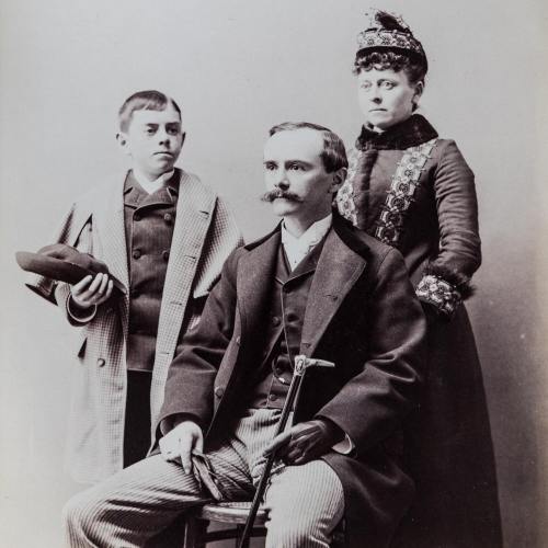 A vintage black-and-white photo of a man seated with a cane, flanked by a boy and a woman in formal attire.