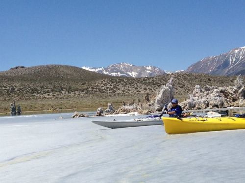 A kayaker in a yellow kayak paddles on a serene lake with rocky formations and snow-capped mountains in the background.