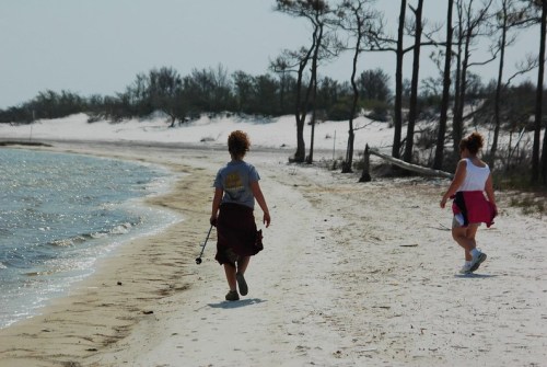 Two women walk along a sandy beach, with trees in the background and water gently lapping at the shore.