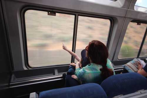 A woman sits by a train window, pointing outside while another passenger relaxes nearby.