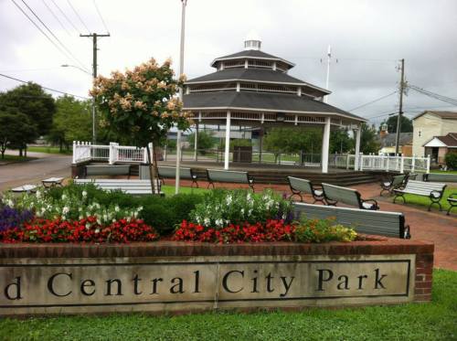 A gazebo in a park surrounded by colorful flowers and benches, with a cloudy sky in the background.