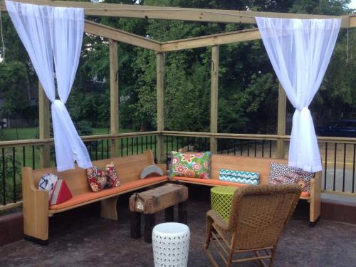 A cozy outdoor seating area with wooden benches, colorful cushions, and sheer curtains, surrounded by greenery.
