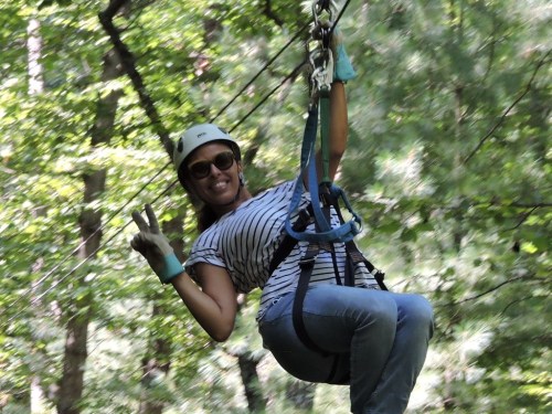 A woman in a helmet and sunglasses smiles and poses with a peace sign while zip-lining through a forest.