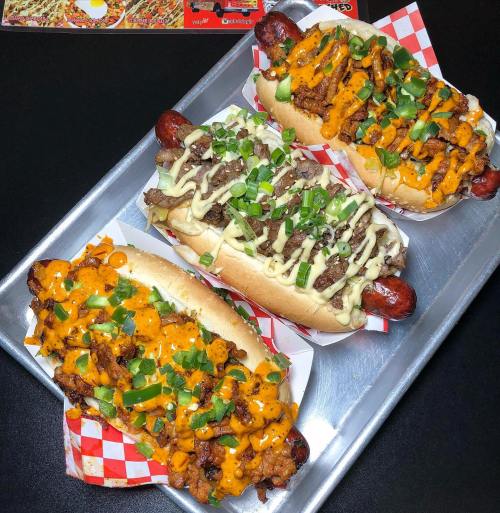 Three gourmet hot dogs on a tray, topped with various sauces, green onions, and colorful ingredients on a checkered paper.
