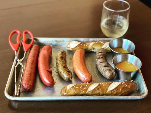 A metal tray with various sausages, pretzel buns, and two small bowls of mustard, alongside a glass of white wine.