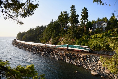 A train curves along a rocky coastline, surrounded by trees and a large house on a hillside.