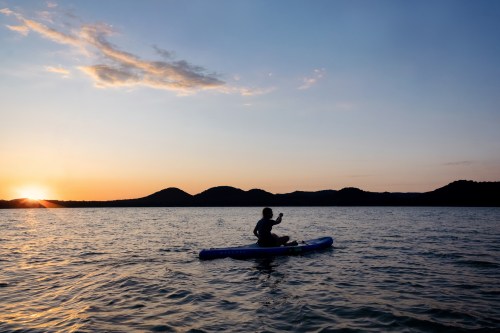 A person kayaking on a calm lake at sunset, with mountains silhouetted in the background.