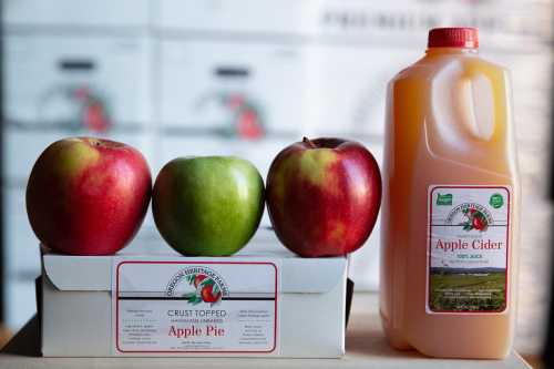 Three apples (red and green) sit beside a carton of apple cider and a box labeled "Crust Topped Apple Pie."