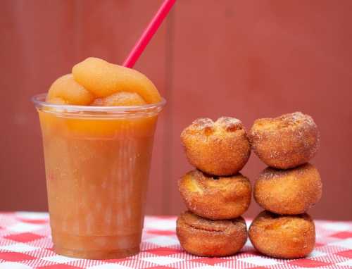 A cup of slushy drink with a straw next to a stack of five golden-brown mini donuts on a checkered tablecloth.