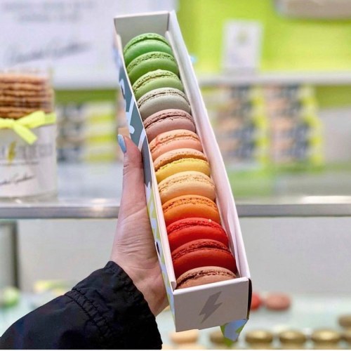 A hand holds a box of colorful macarons arranged in a gradient from green to red, set against a bright background.