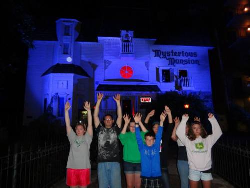 A group of six people with hands raised in front of a spooky, illuminated mansion labeled "Mysterious Mansion."
