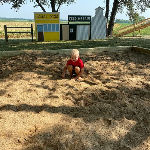 A young child in a red shirt plays in a sandy area with colorful wooden buildings in the background.