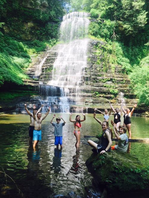A group of people joyfully poses in a river near a waterfall, surrounded by lush greenery on a sunny day.