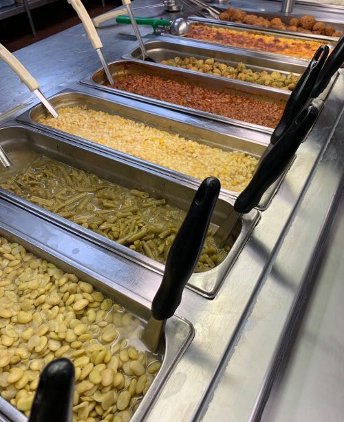 A buffet line featuring various trays of colorful side dishes, including beans, corn, and rice.