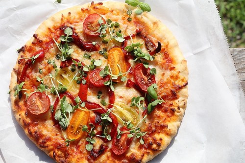 A freshly baked pizza topped with colorful tomatoes, bell peppers, and microgreens on a white background.