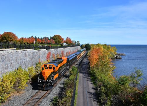 A colorful train travels along a scenic route by a lake, surrounded by autumn foliage and clear blue skies.