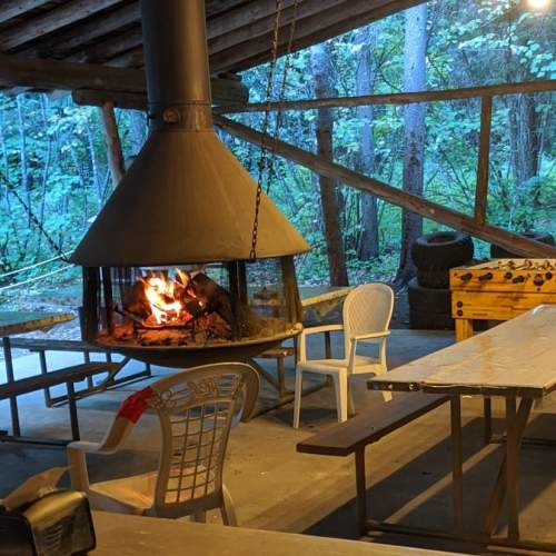 A cozy outdoor shelter with a central fireplace, surrounded by tables and chairs, set in a wooded area.