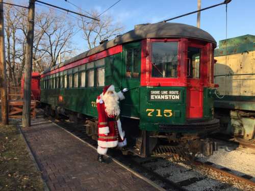 Santa Claus stands beside a vintage green and red train car labeled "715" at a railway station.