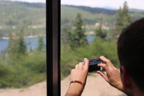 A person takes a photo of a scenic view through a window, surrounded by trees and a lake in the background.