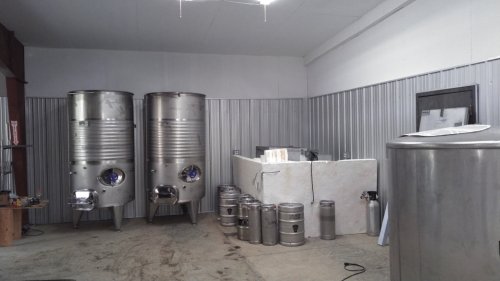 Interior of a brewery featuring stainless steel fermentation tanks and storage kegs against a metal-paneled wall.