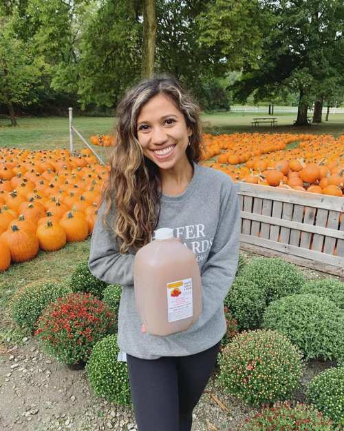 A smiling woman holds a jug of cider in a pumpkin patch, surrounded by orange pumpkins and green bushes.