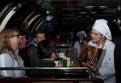 A chef in a white coat engages with a group of diners on a festive train, sharing a menu or story.