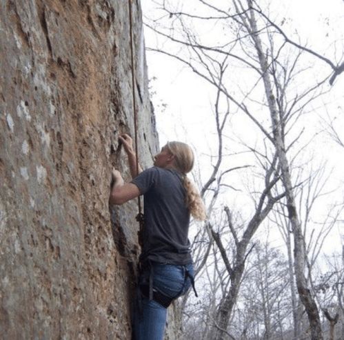 A climber ascends a rocky wall surrounded by bare trees on a cloudy day.
