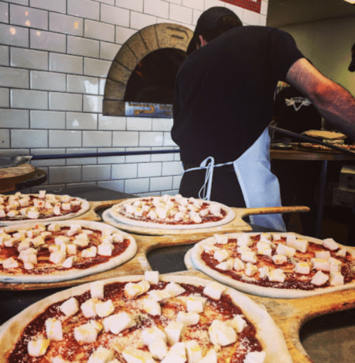 A chef prepares pizzas topped with cheese in a kitchen, with an oven in the background.